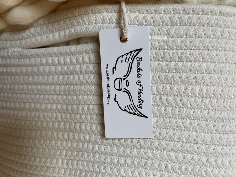 A Baskets of Healing tag with the winged-basket logo tied to the handle of a cream rope basket