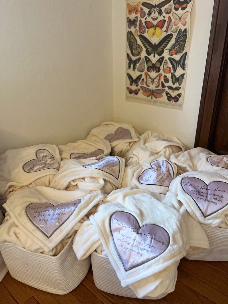 Several finished Baskets of Healing lined up in a corner, each topped with a cream blanket embroidered with a heart